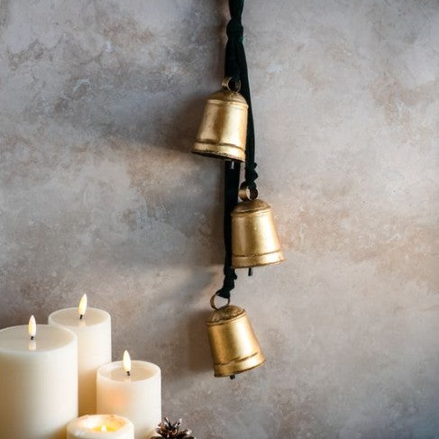 Three lit candles with a decorative hanging brass bell on a textured wall.