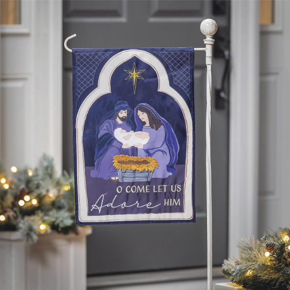 Decorative Christmas flag with nativity scene and 'O Come Let Us Adore Him' text, displayed outdoors.