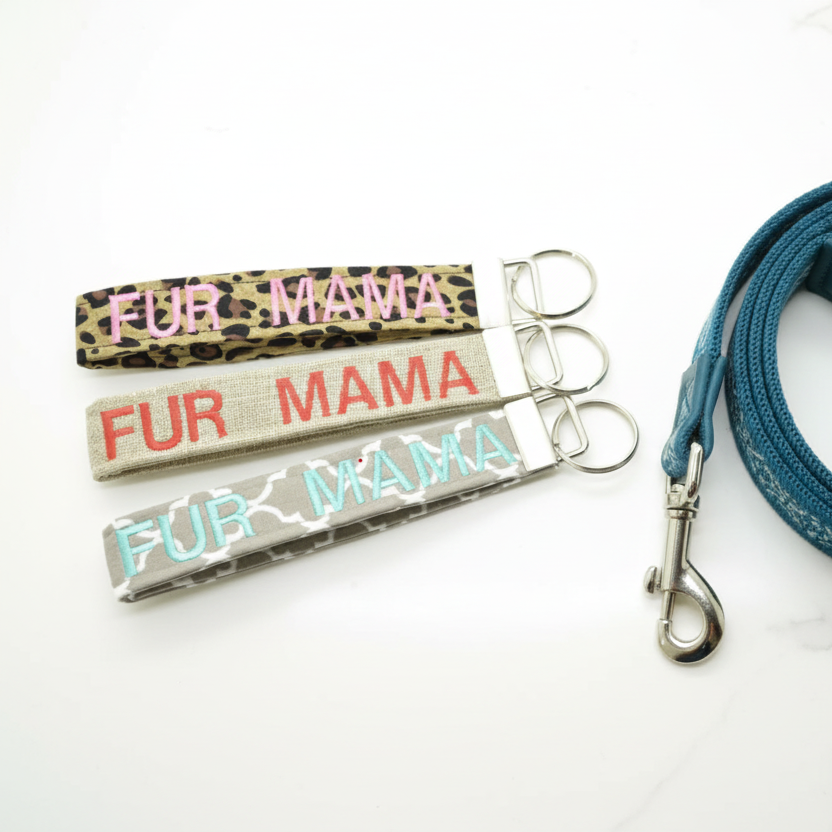 Three keychains embroidered with FUR MAMA on leopard print, burlap style, and quatrefoil fabric on a countertop next to a leash.