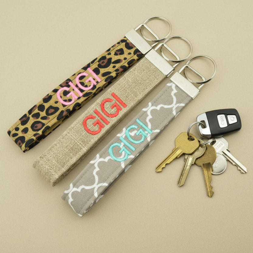 Three fabric keychains embroidered with 'GIGI'. Patterns are leopard, burlap, and grey quatrefoil. On a tan background with keys nearby.