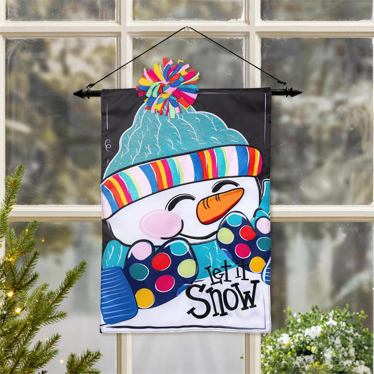 Decorative flag with a colorful snowman design and 'Let It Snow' text, hanging in front of a window.