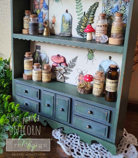 A decorative mold with a green cabinet and a shelf, adorned with floral and natural motifs, including mushrooms and butterflies, and labeled 'The Painted Unicorn'.