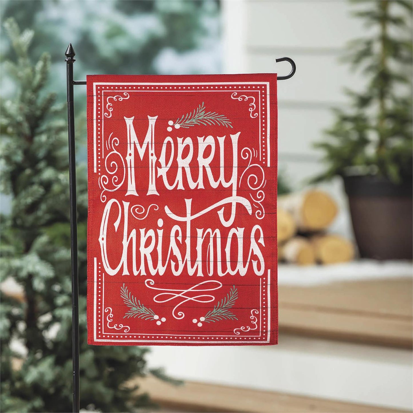 Red 'Merry Christmas' garden flag with decorative elements on a pole against a blurred indoor background.