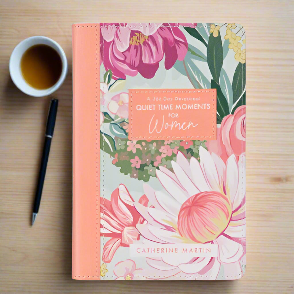 A daily devotional book titled 'Quiet Time Moments for Women' by Catherine Martin, featuring a floral design cover in shades of pink, peach, and green with a peach-colored faux leather spine. Accompanied by a black pen and a cup of tea on a wooden surface.