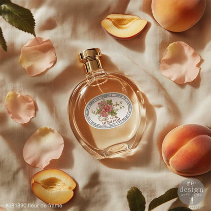 Perfume bottle with Fleur De Frame Luxe Transfers surrounded by peach slices and petals on a textured surface