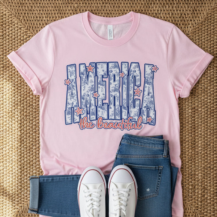 Pink tshirt with "America the beautiful" graphic design on a rattan background with jeans and white sneakers.