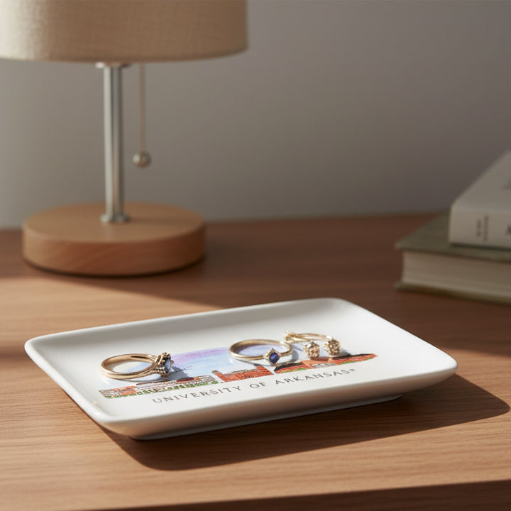 Rectangular white plate with a colorful illustration of a building and 'University of Arkansas' text sitting on a nighstand with a few rings on it