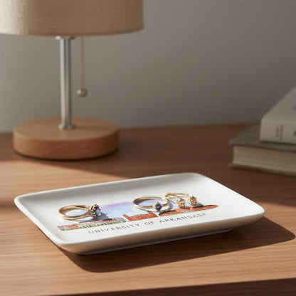 Rectangular white plate with a colorful illustration of a building and 'University of Arkansas' text sitting on a nighstand with a few rings on it