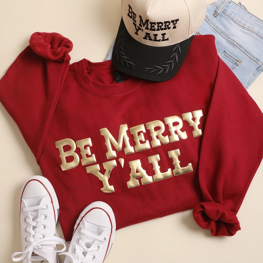 Red sweatshirt with 'Be Merry Y'all' text, white sneakers, and a cap on a beige background