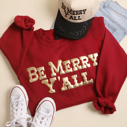 Red sweatshirt with 'Be Merry Y'all' text, white sneakers, and a cap on a beige background