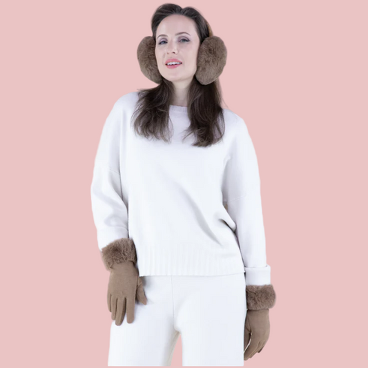 Woman wearing a white sweater, white pants, brown gloves, and brown ear muffs on a pink background
