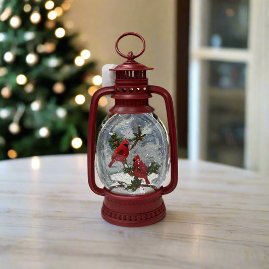 Red lantern-shaped snow globe with cardinals on a white surface with a Christmas tree in the background.