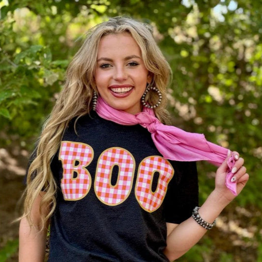 Woman wearing a black t-shirt with 'BOO' in red and yellow checkered letters, holding a pink scarf outdoors.