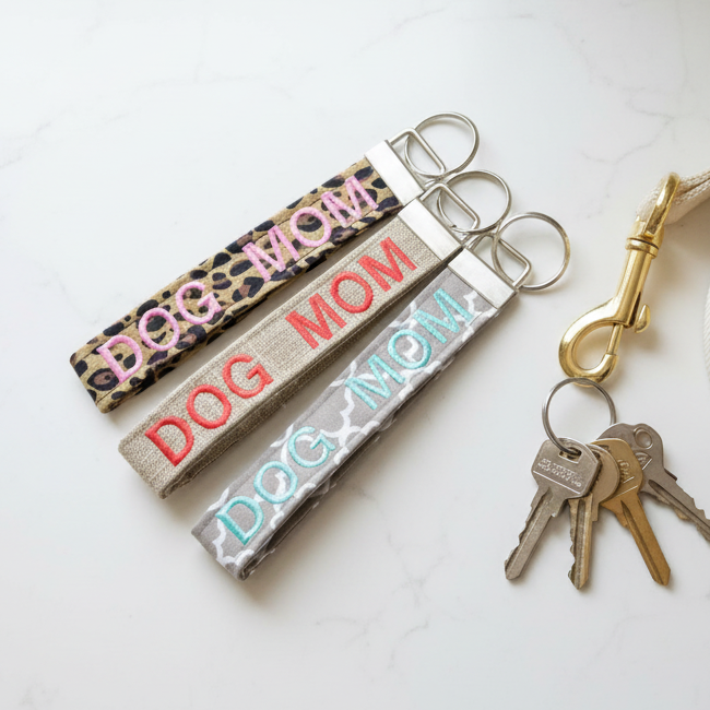 Three keychains embroidered with DOG MOM on leopard print, burlap style, and quatrefoil fabric on a countertop next to keys and a leash.