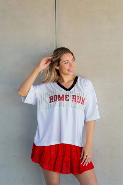 Woman wearing a white oversized sports jersey with 'HOME RUN' printed on it, standing against a plain wall.