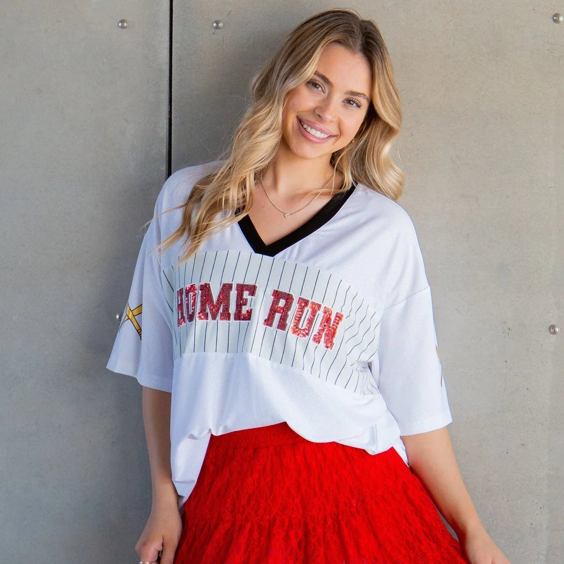 Woman wearing a white oversized sports jersey with 'HOME RUN' text and a red skirt against a gray wall.