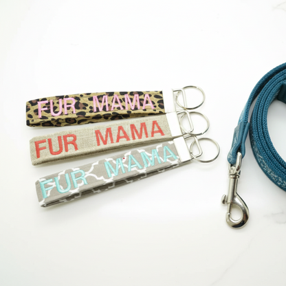 Three keychains embroidered with FUR MAMA on leopard print, burlap style, and quatrefoil fabric on a countertop next to a leash.