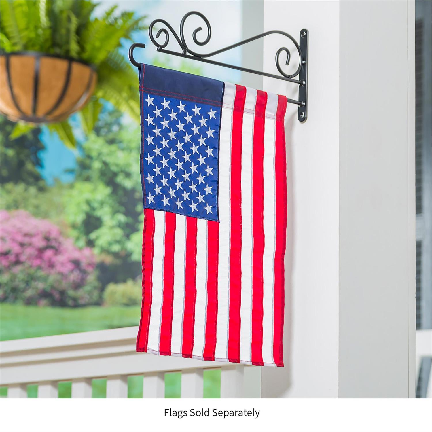 American flag hanging on a decorative metal wall hook with a garden and building in the background.