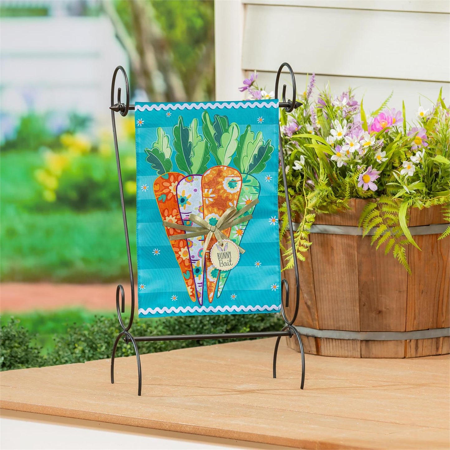 Evergreen Bunny Bait Bunch garden flag with carrots on a stand next to a wooden planter with flowers