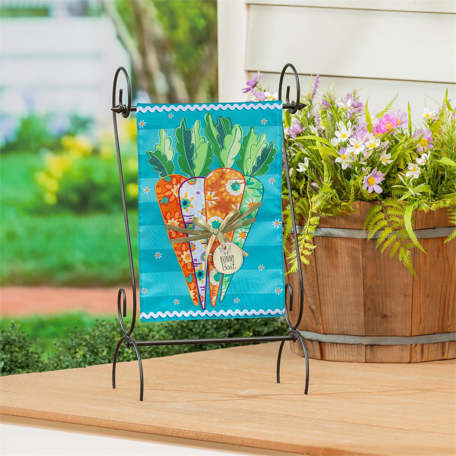 Evergreen Bunny Bait Bunch garden flag with carrots on a stand next to a wooden planter with flowers