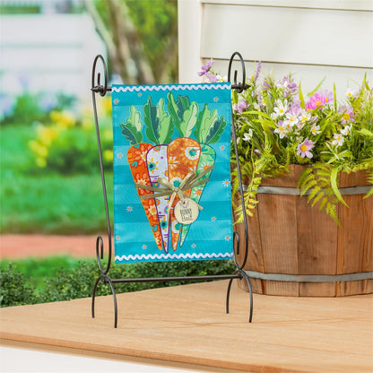 Evergreen Bunny Bait Bunch garden flag with carrots on a stand next to a wooden planter with flowers