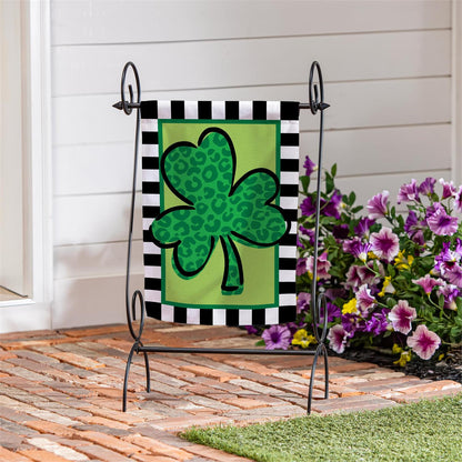 Evergreen Patterned Shamrock suede garden flag with a green shamrock with a black and white striped frame on a stand, placed outdoors.