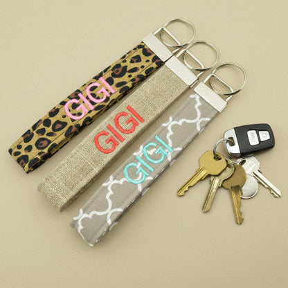 Three fabric keychains embroidered with 'GIGI'. Patterns are leopard, burlap, and grey quatrefoil. On a tan background with keys nearby.