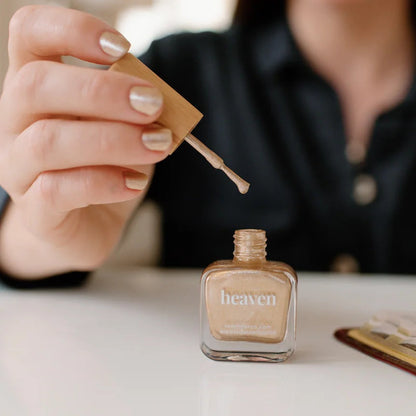 Person applying gold nail polish from a bottle labeled 'heaven' with a blurred background.