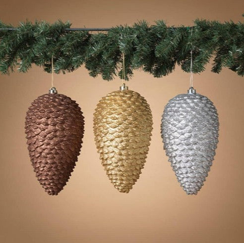 Three pine cone-shaped ornaments in bronze, gold, and silver hanging against a beige background.