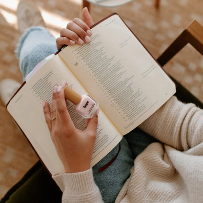 Woman holding a bottle of Reminder Nail Polish in Hope Light Pink with a Bible on her lap.