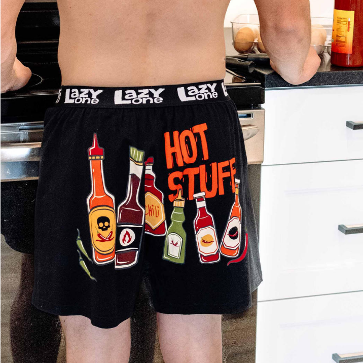 Person wearing black boxer briefs with hot sauce bottle design and 'Lazy One' waistband in a kitchen.