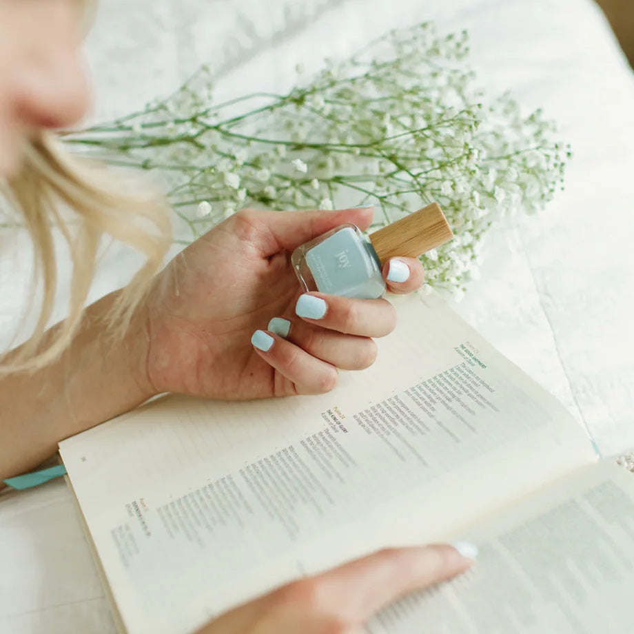 A woman reading the Bible holding a bottle of Reminder Nail Polish in Joy Light Blue.