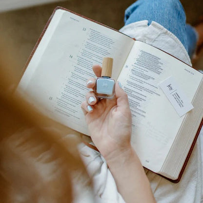 Person holding aa bottle of Joy light blue nail polish over an open Bible and showing the devotion card.