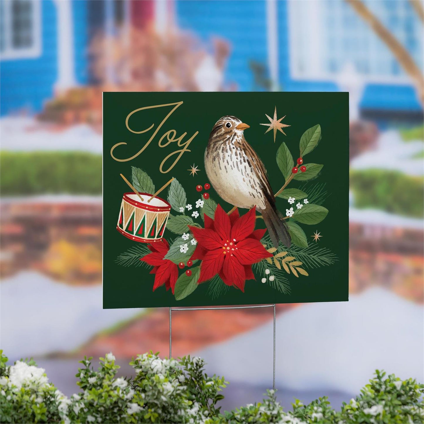 Christmas-themed yard sign with a bird, drum, and poinsettia in front of a blurred house and pond background.
