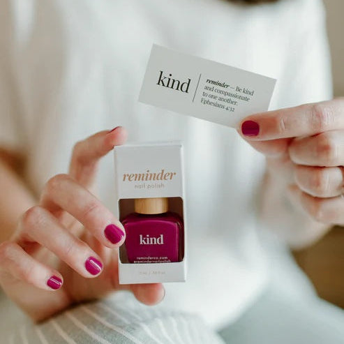 Person holding a  bottle of Reminder Nail Polish in Kind Berry Magenta and devotion card.