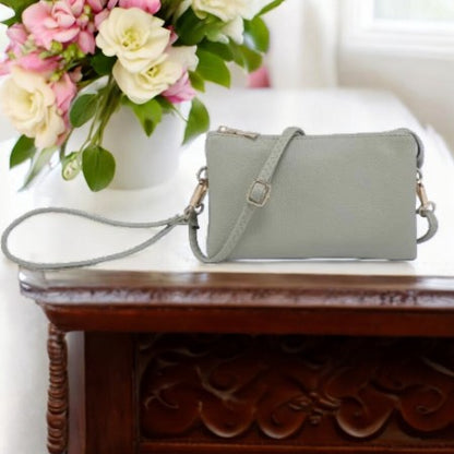 LIGHT SAGE GREEN HANDBAG ON A BROWN TABLE WITH A PINK AND WHITE FLORAL ARRANGEMENT IN THE BACKGROUND