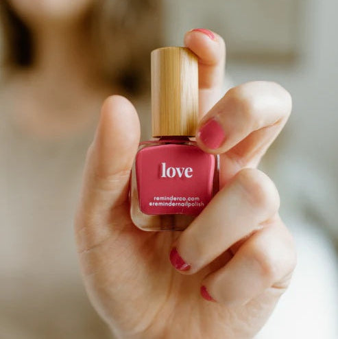 Hand holding a bottle of Reminder Nail Polish in Love Watermelon Red, blurred background