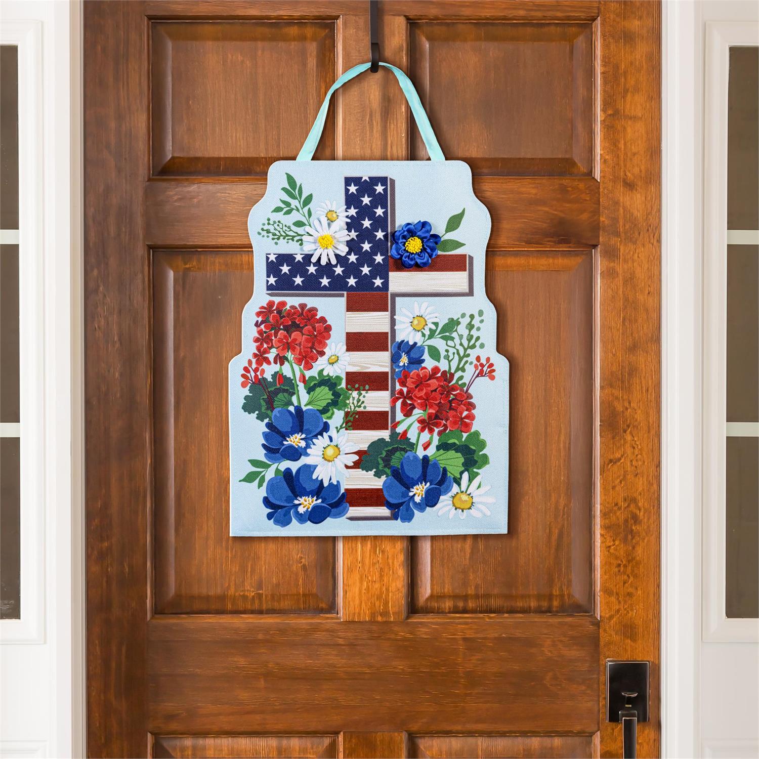 Patriotic Cross door hanger designed  with stars and stripes and flowers hanging on a wooden door.