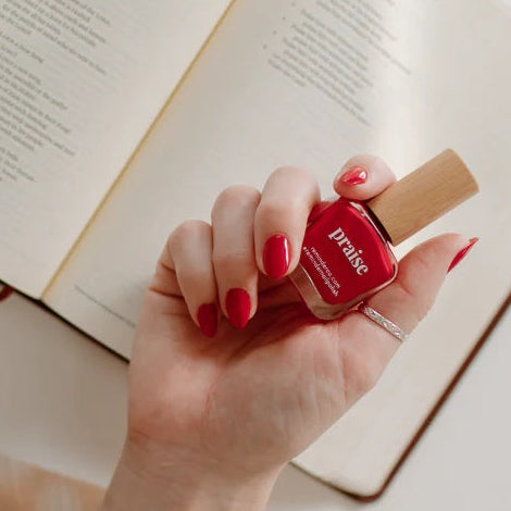 Hand holding a bottle of  Reminder Nail Polish in Praise Classic Red against the background of a blurred Bible.