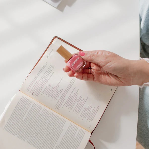 Hand holding a bottle of Reminder Nail Polish in Purpose Honeysuckle Pink above an open Bible.