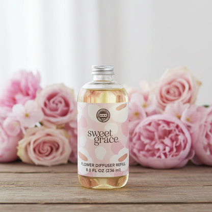 Bottle of Sweet Grace Diffuser Oil Refill on a wood table with pink flowers in the background