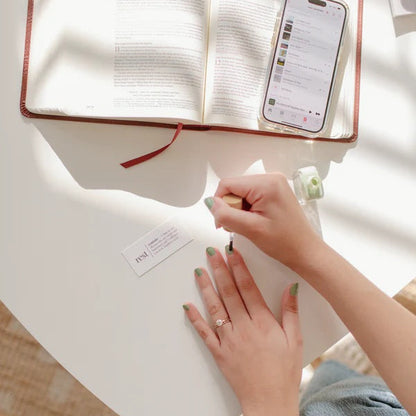 Woman painting her nails with Reminder Nail Polish in Rest Sage Green on a table with an open Bible and cell phone.