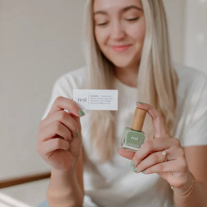 Woman holding a bottle of Reminder Nail Polish in Rest Sage Green and a devotion card.