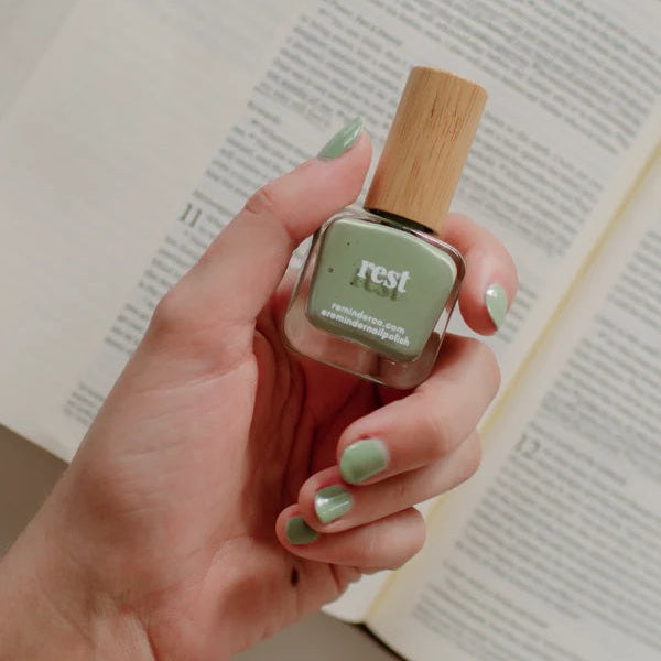Hand holding a bottle of 'Reminder Nail Polish in Rest Sage Green over a blurred open Bible.