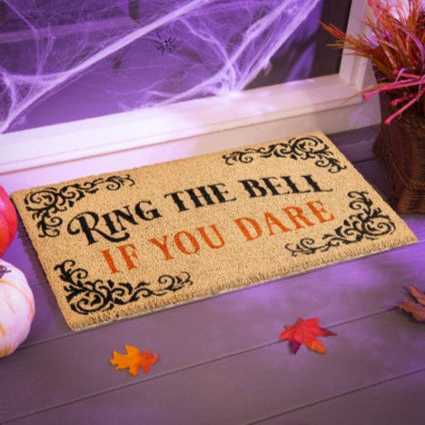 Decorative doormat with Halloween text on a wooden floor with pumpkins and leaves.