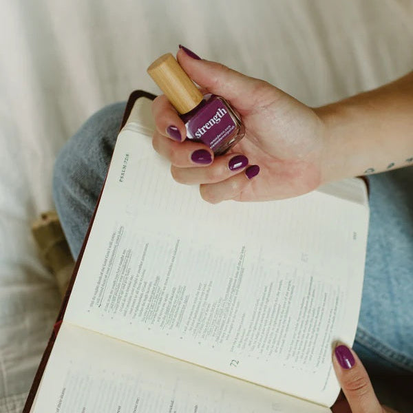 Hand holding a bottle Reminder Nail Polish in Violet Purple over a Bible.