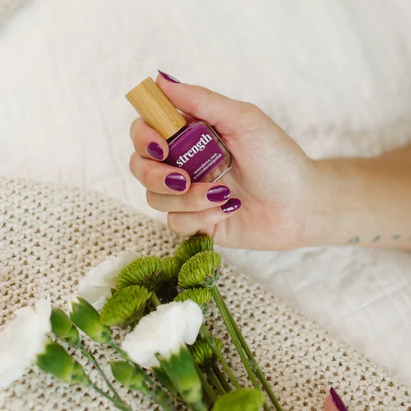 Hand holding a bottle of Reminder Nail Polish in Violet Purple with flowers in the foreground